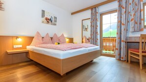 A bedroom with a wooden bed, bedside lamps, a painting, and a view through a window with curtains. - Apartment "Panorama" with Mountain Views, Shared Garden, and Wi-Fi (Mittersill)