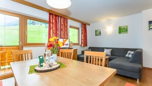 A modern living room with a wooden dining table, a grey sofa, and large windows with red curtains. - Apartment "Panorama" with Mountain Views, Shared Garden, and Wi-Fi (Mittersill)