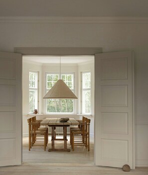 Un comedor con mesa y sillas de madera, una lámpara colgante y ventanas amplias que dan a zonas verdes.