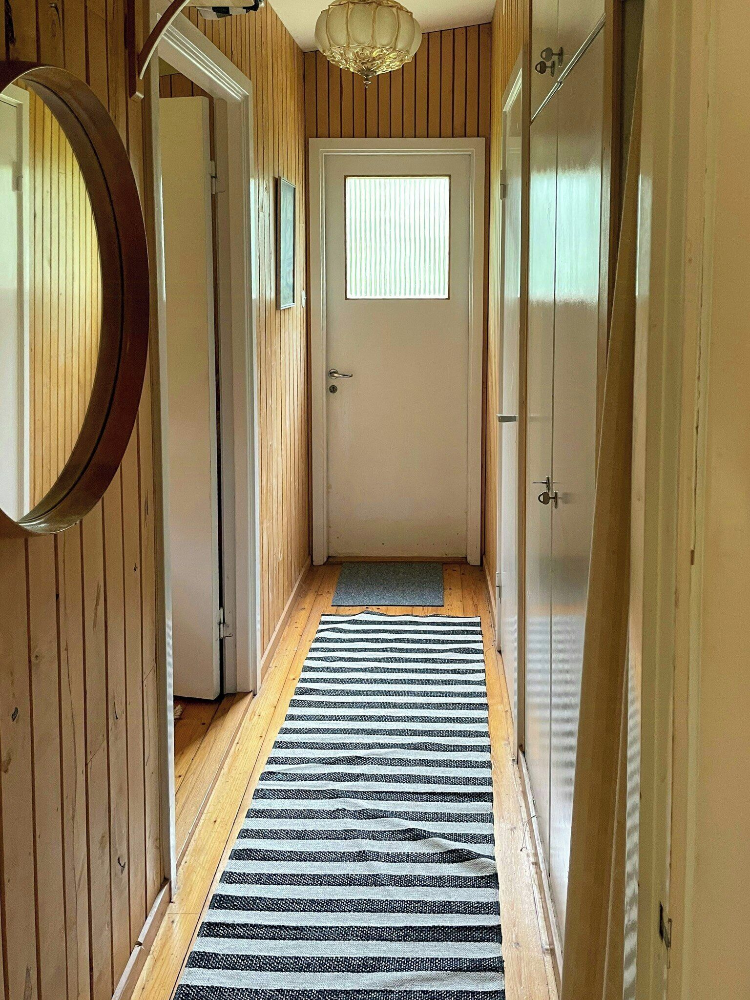 A hallway with wooden walls, a door with a window, a mirror, and a striped rug.