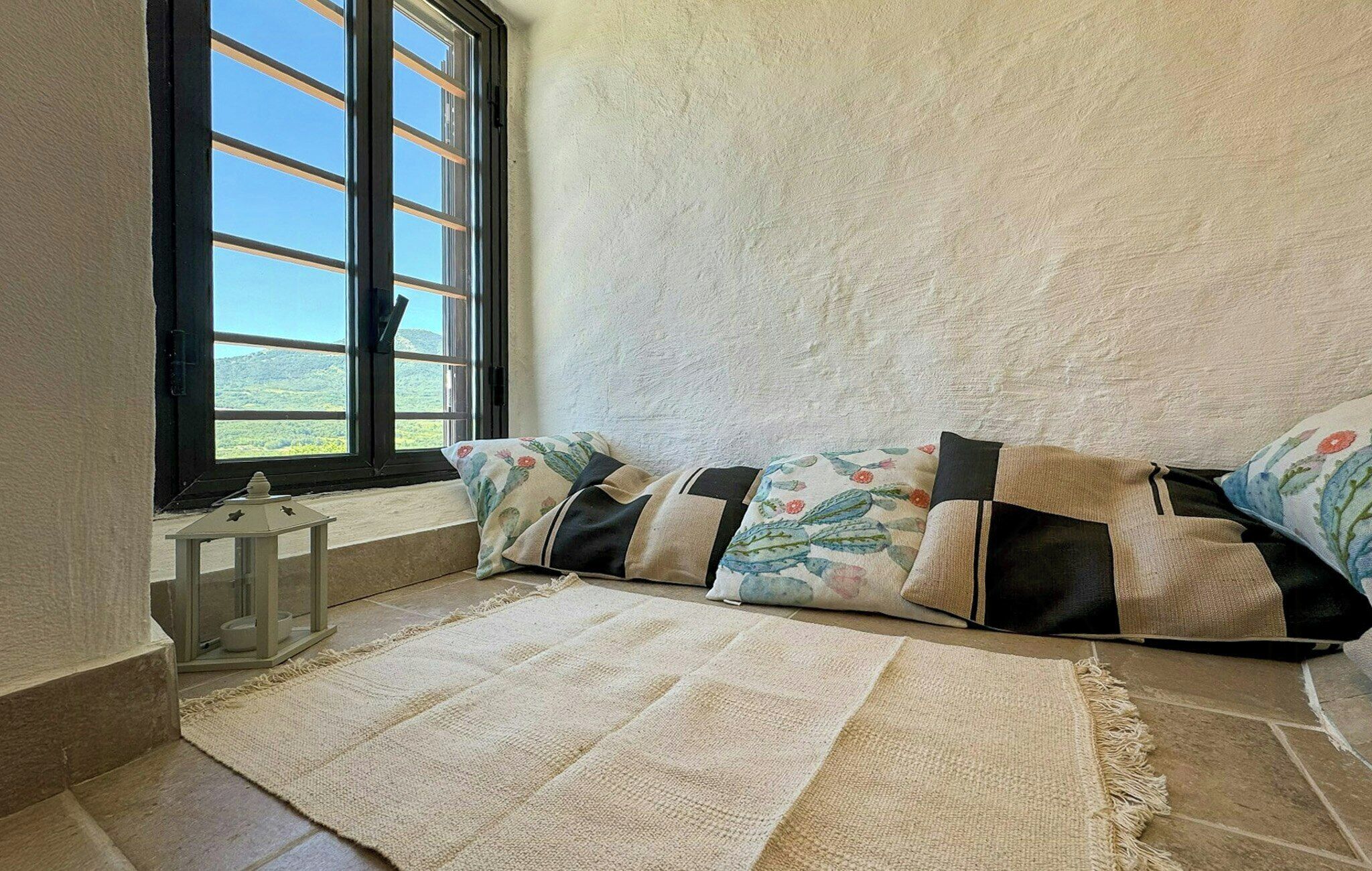 A room with a large window, a cushioned bench, a small table, and a rug.