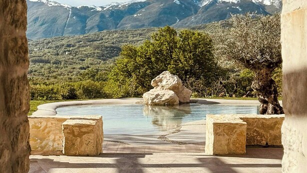 Una piscina con montañas al fondo, rodeada de construcciones de piedra y vegetación.