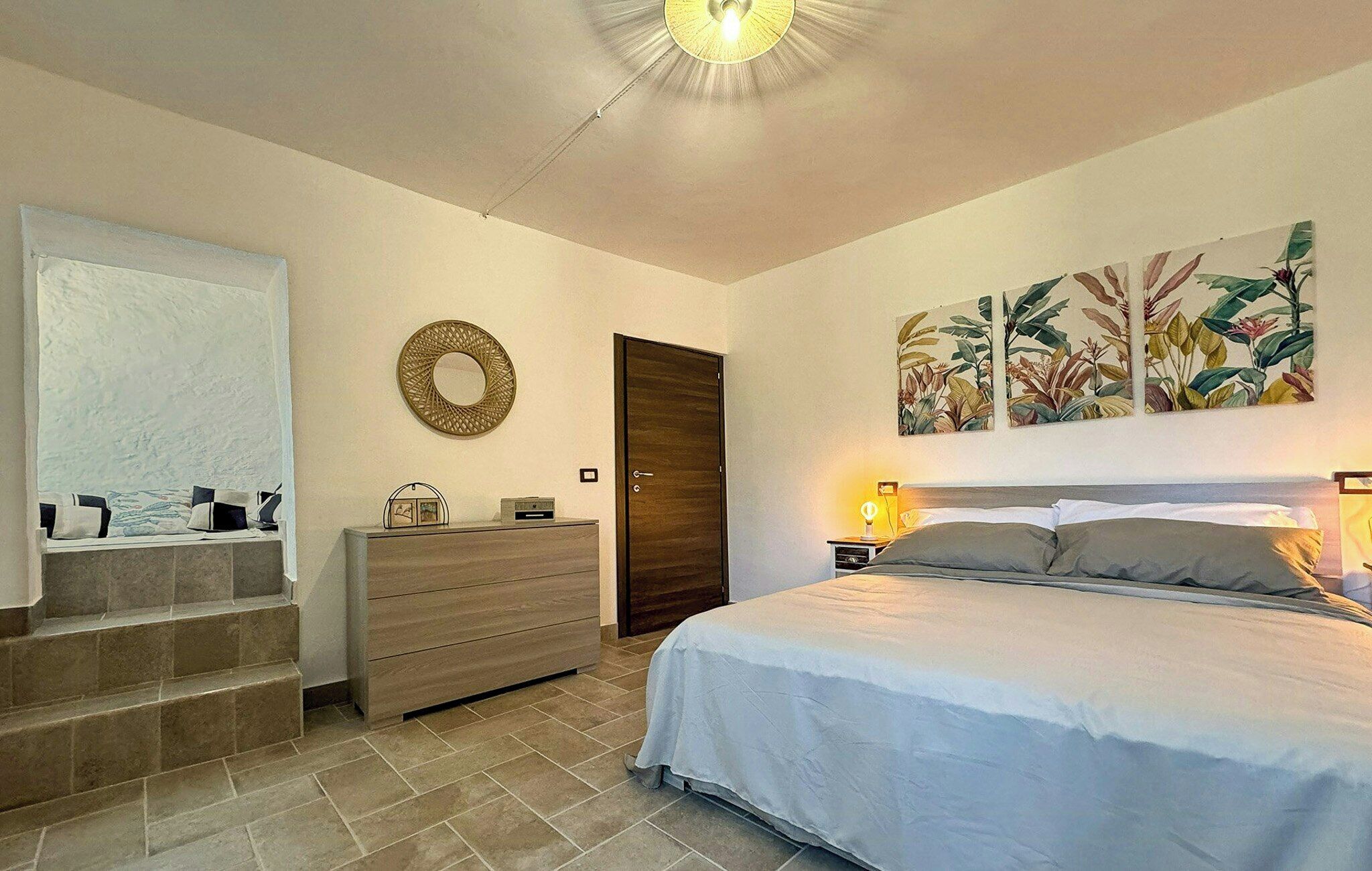 A bedroom with a bed, a dresser, a mirror, a door, and a wall with a plant painting.