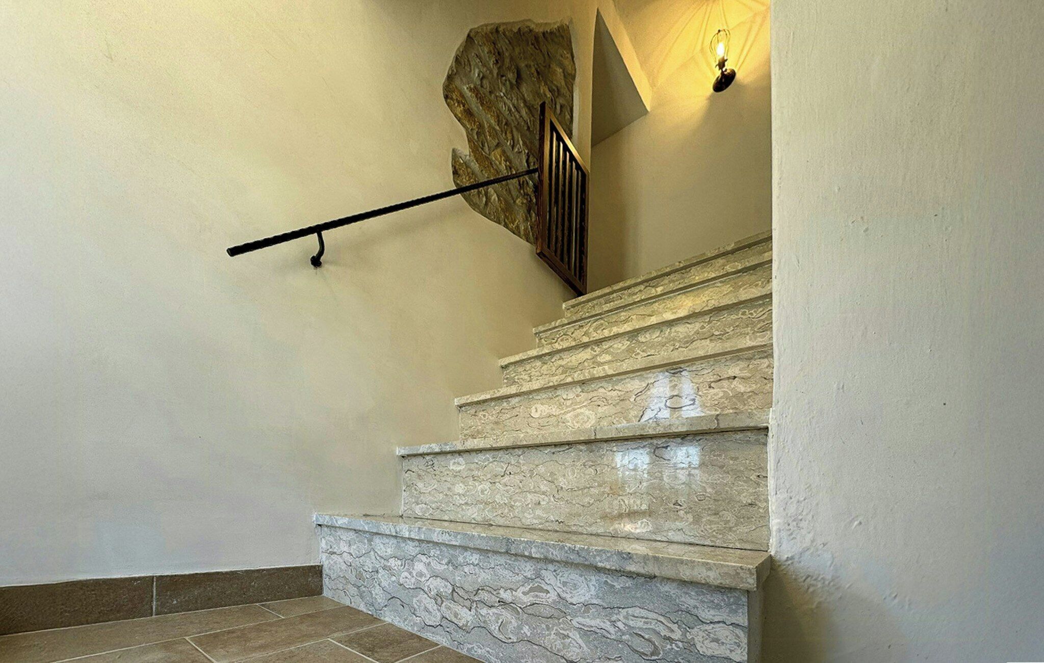 A staircase with stone steps and a handrail, a wall with a textured pattern, and a light fixture.