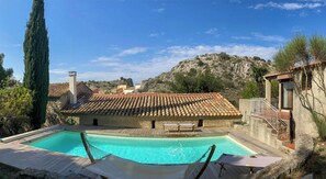 Ein Pool mit einer Hängematte und Liegestühlen, ein Steinhaus mit einem Ziegeldach und eine gebirgige Landschaft im Hintergrund.