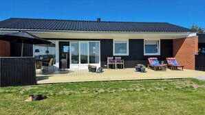 Ein Haus mit überdachter Terrasse, zwei Liegestühlen und einem Hund im Garten.