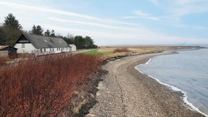 A coastal building with a gravel path leading to the sea, surrounded by vegetation and a clear sky.