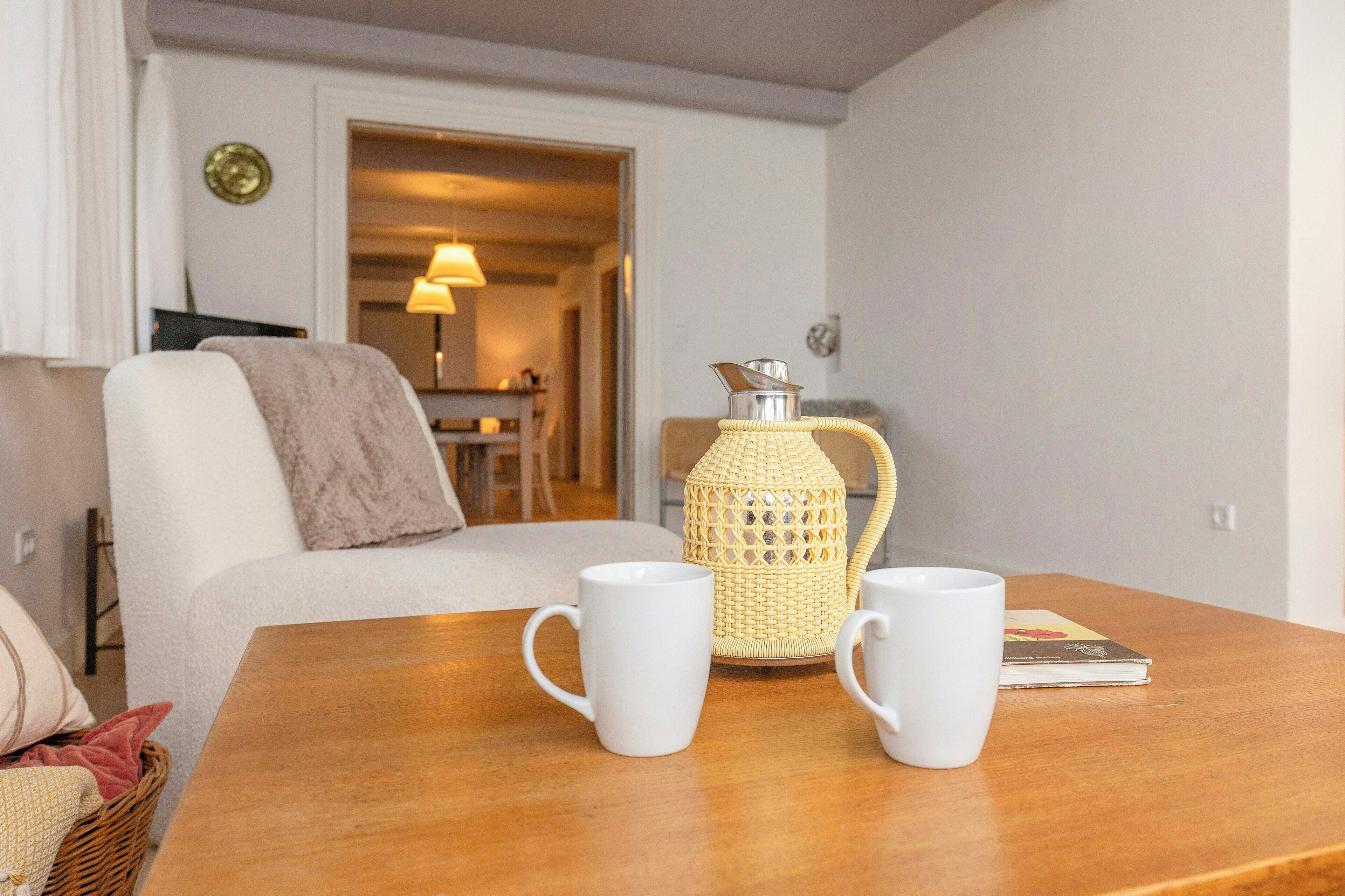A wooden table with two white mugs, a woven lantern, and a book.