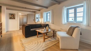 A spacious living room with a sofa, armchair, coffee table, and a rug. There are two windows with curtains, a door leading to another room, and a painting on the wall.