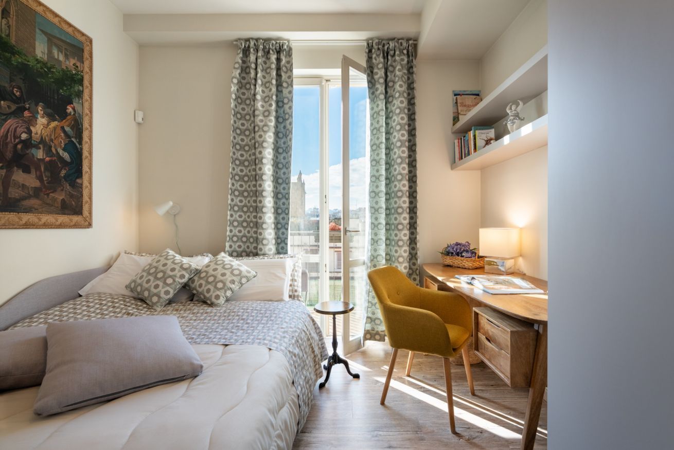 A bedroom with a bed, a desk, a chair, and a view through a window.