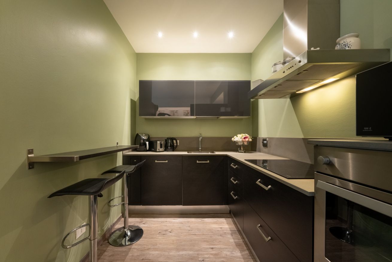 A compact kitchen with dark cabinetry, a stainless steel oven, and a single bar stool.
