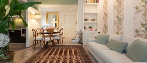 Ein Wohnzimmer mit einer Couch, einem Esstisch mit Stühlen, einem gemusterten Teppich und einem Bücherregal mit Dekoration.