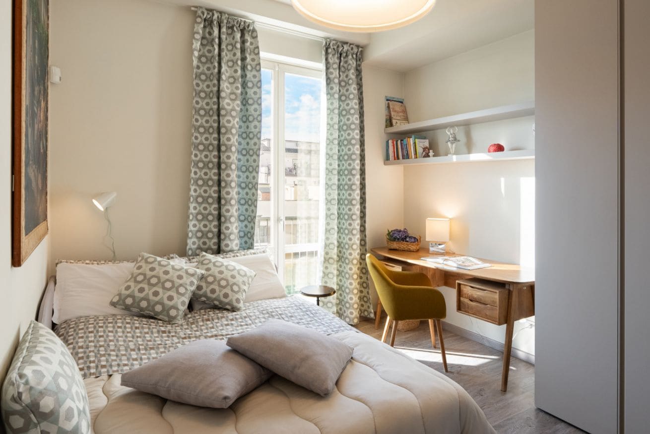 A bedroom with a bed, a desk, a chair, a lamp, and a window with curtains.