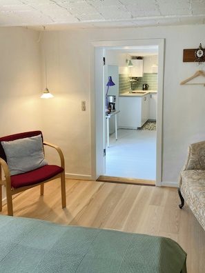 A room with a wooden floor, a red chair with a pillow, a green blanket, and a doorway leading to a kitchen area.