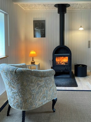 A room with a patterned armchair, a small table with a lamp, a fireplace with a black chimney, and a framed quote on the wall.
