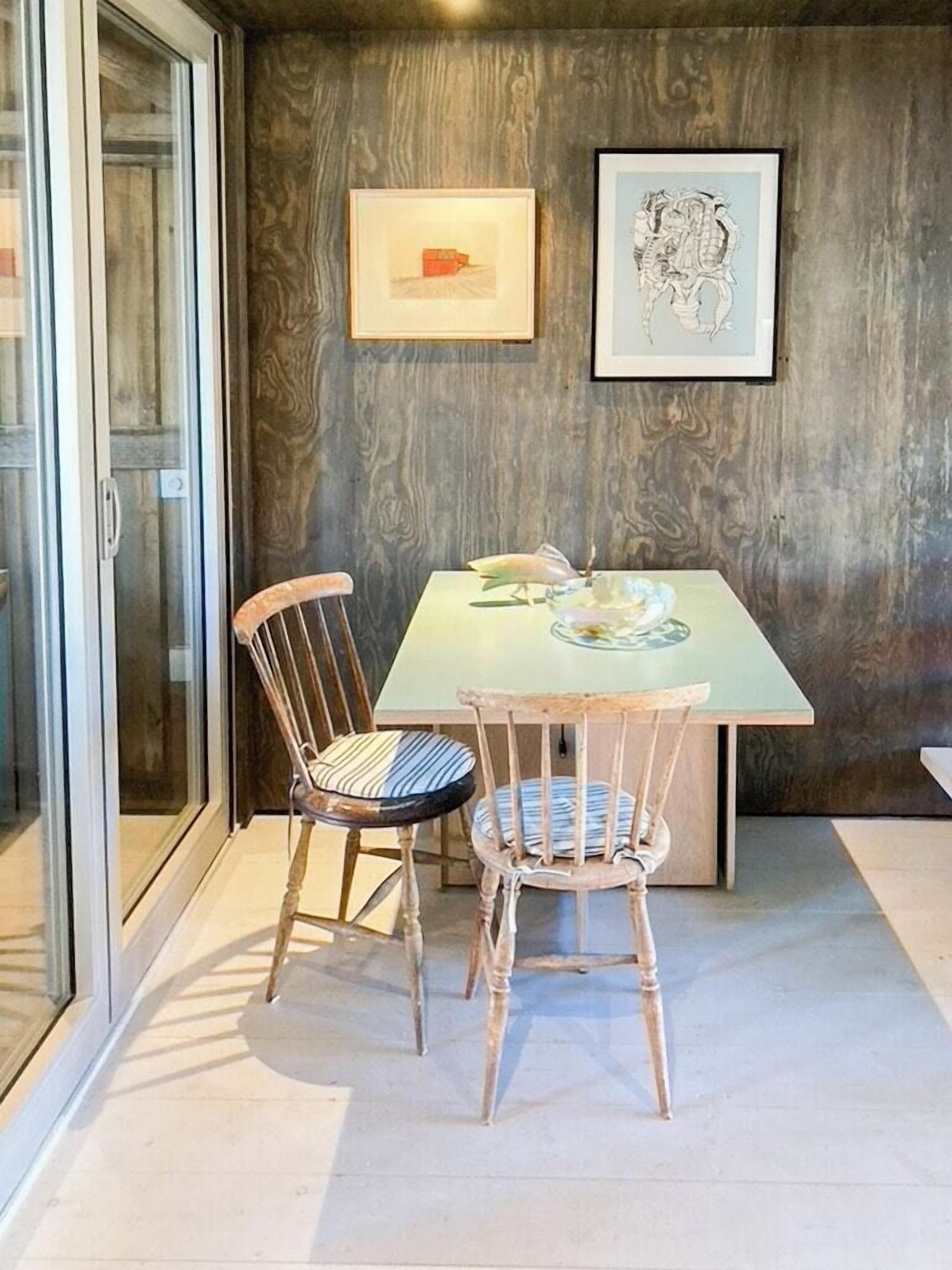 Ein Essbereich mit Glastür, zwei Stühlen, einem Tisch und zwei gerahmten Kunstwerken an der Wand.