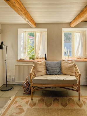 Ett rum med en soffa i rotting, två fönster med gardiner, ett element och en golvlampa.