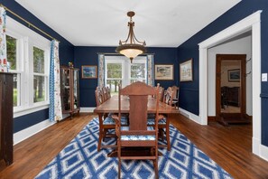 A dining room with a large wooden table, chairs, a chandelier, and a patterned rug.