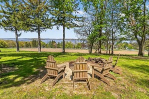 Ein hölzerner Feuerkorb, umgeben von Adirondack-Stühlen, in einer grünen Wiese mit Bäumen und einem See im Hintergrund.