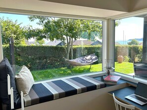 Ein Zimmer mit einem großen Fenster, einer gepolsterten Bank, einem Laptop auf einem Tisch und einer Hängematte davor.
