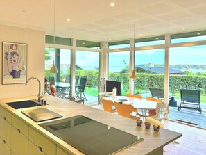 Eine moderne Küche mit einer großen Kochinsel, einem Essbereich mit orangefarbenen Stühlen und großen Fenstern mit Blick ins Freie.