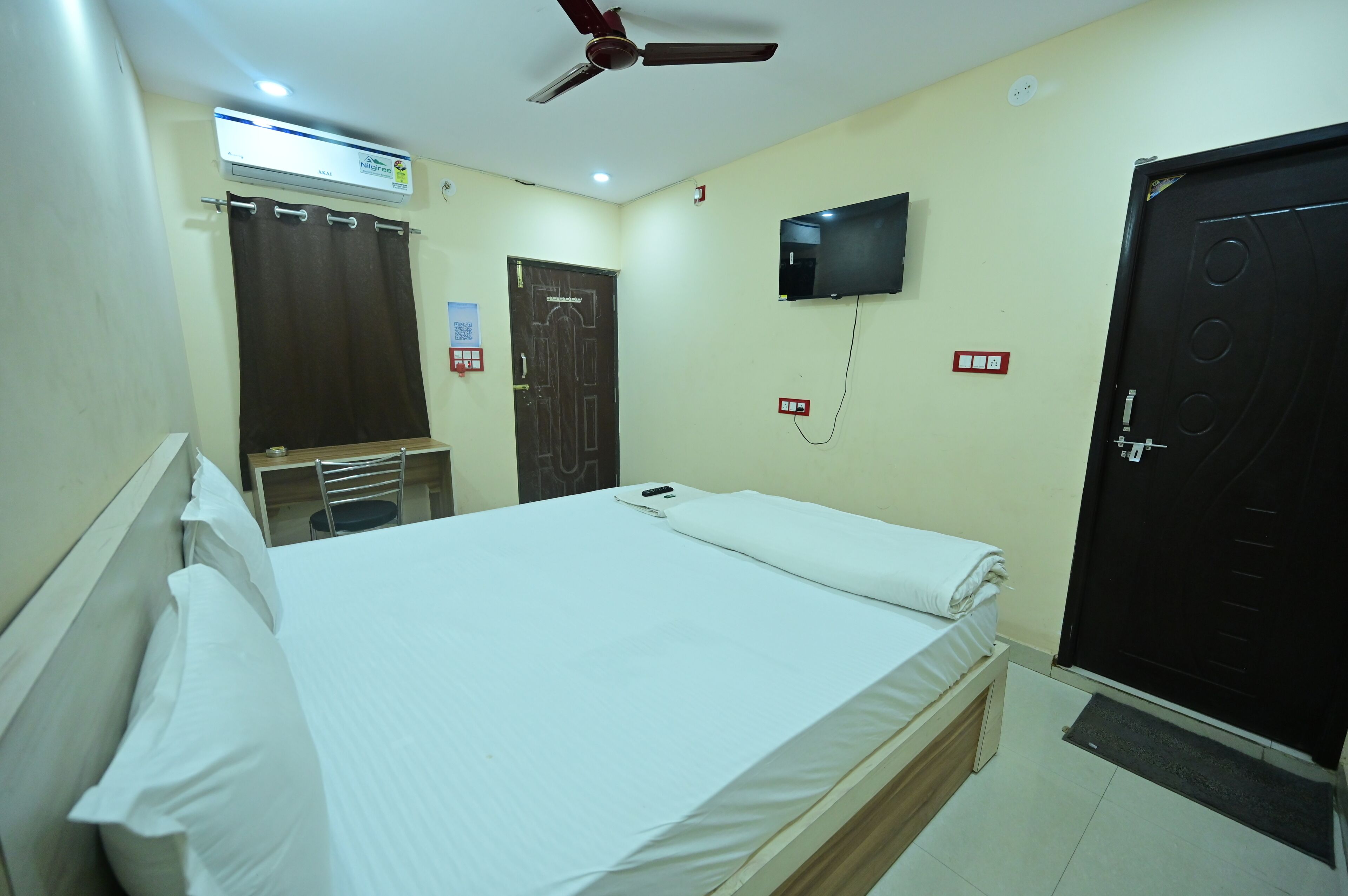 Habitación de hotel con una cama, ventilador de techo, televisor montado en la pared, una puerta y una ventana.