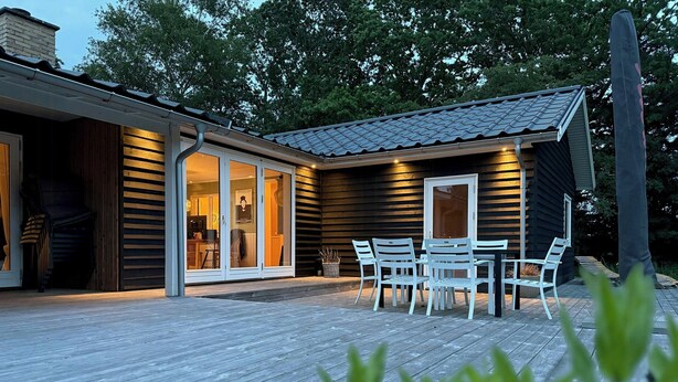 Eine Holzterrasse mit Sitzgelegenheiten im Freien, ein Glastür-Eingang und ein Gebäude mit dunklen Fassadenplatten und einem Ziegeldach.