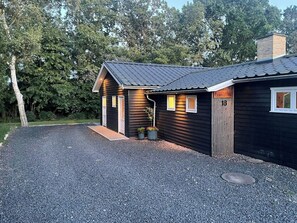 A small, dark wooden house with a gravel driveway and a metal roof. - Modern family house near beach in Odsherred (Vig)