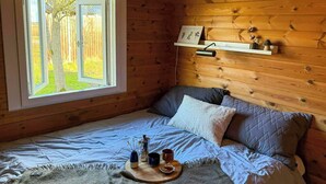A wooden cabin room with a bed, a window with a view of trees, a shelf with decorative items, and a bedside table with a cup and a vase. - Log cabin with water views near Fjellerup beach (Ørsted)