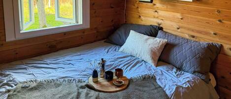 Quarto de cabana de madeira com uma cama, uma janela com vista para árvores, uma prateleira com objetos decorativos e uma mesa de cabeceira com uma xícara e um vaso.