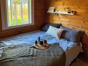 Ein Holzhüttenraum mit einem Bett, einem Fenster mit Blick auf Bäume, einer Ablage mit Dekoration und einem Nachttisch mit Tasse und Vase.