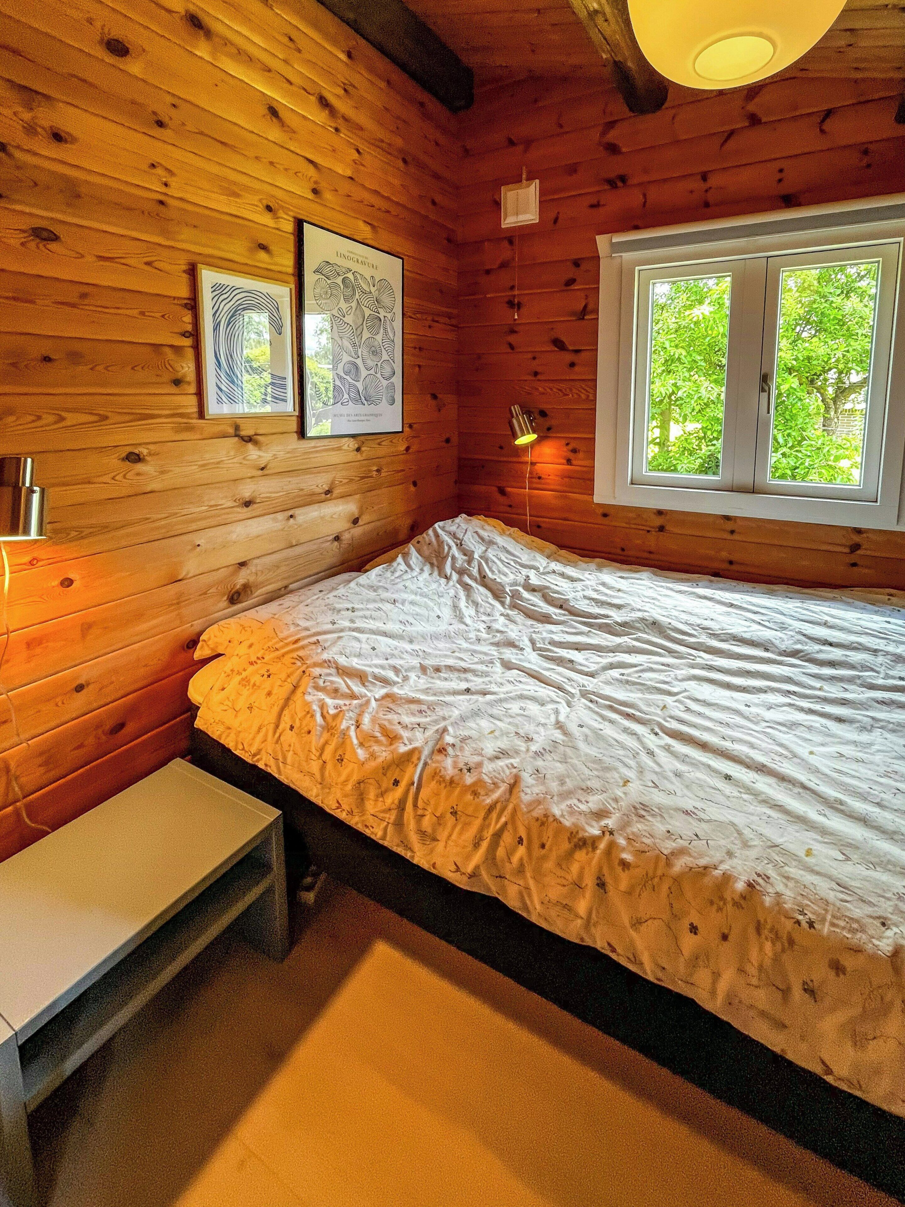 Una habitación de cabaña de madera con una cama, una mesita, dos cuadros enmarcados, una ventana y una lámpara fijada a la pared.