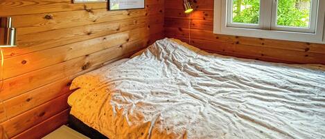 Una habitación de cabaña de madera con una cama, una mesita, dos cuadros enmarcados, una ventana y una lámpara fijada a la pared.