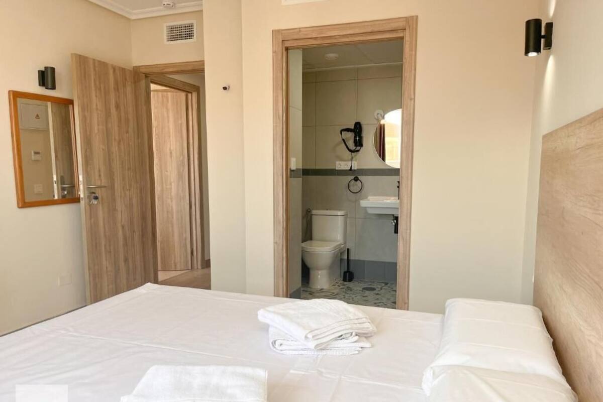 Habitación de hotel con cama, puerta de madera, baño visible a través de una puerta abierta y un espejo en la pared.