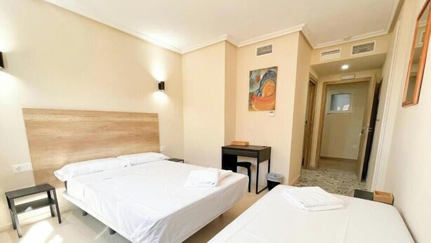 A hotel room with two beds, a desk, a chair, a painting on the wall, and a door leading to another room. - HOTEL LA MARINA (Elche)