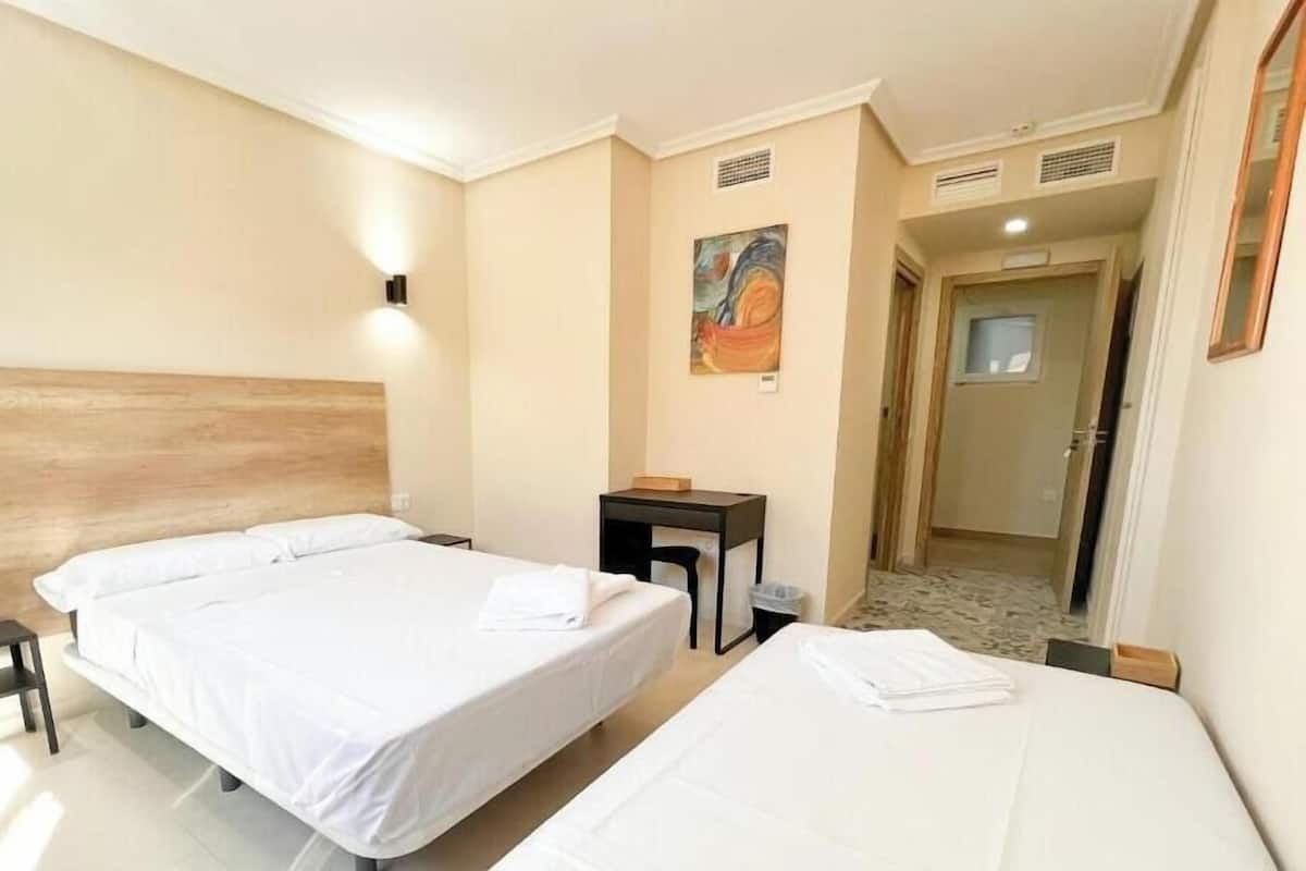 Habitación de hotel con dos camas, un escritorio, una silla, un cuadro en la pared y una puerta que da a otra habitación.