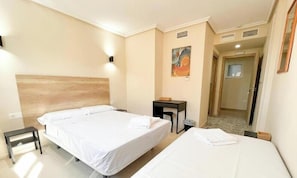 A hotel room with two beds, a desk, a chair, a painting on the wall, and a door leading to another room.