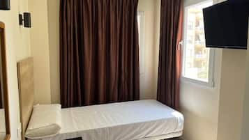 Una cama individual con colchón y almohada blancos, un televisor de pantalla plana montado en la pared, y una ventana con cortinas.