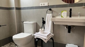 Un baño con inodoro, toallero con toalla, rollo de papel higiénico, lavamanos con dispensador de jabón y espejo.