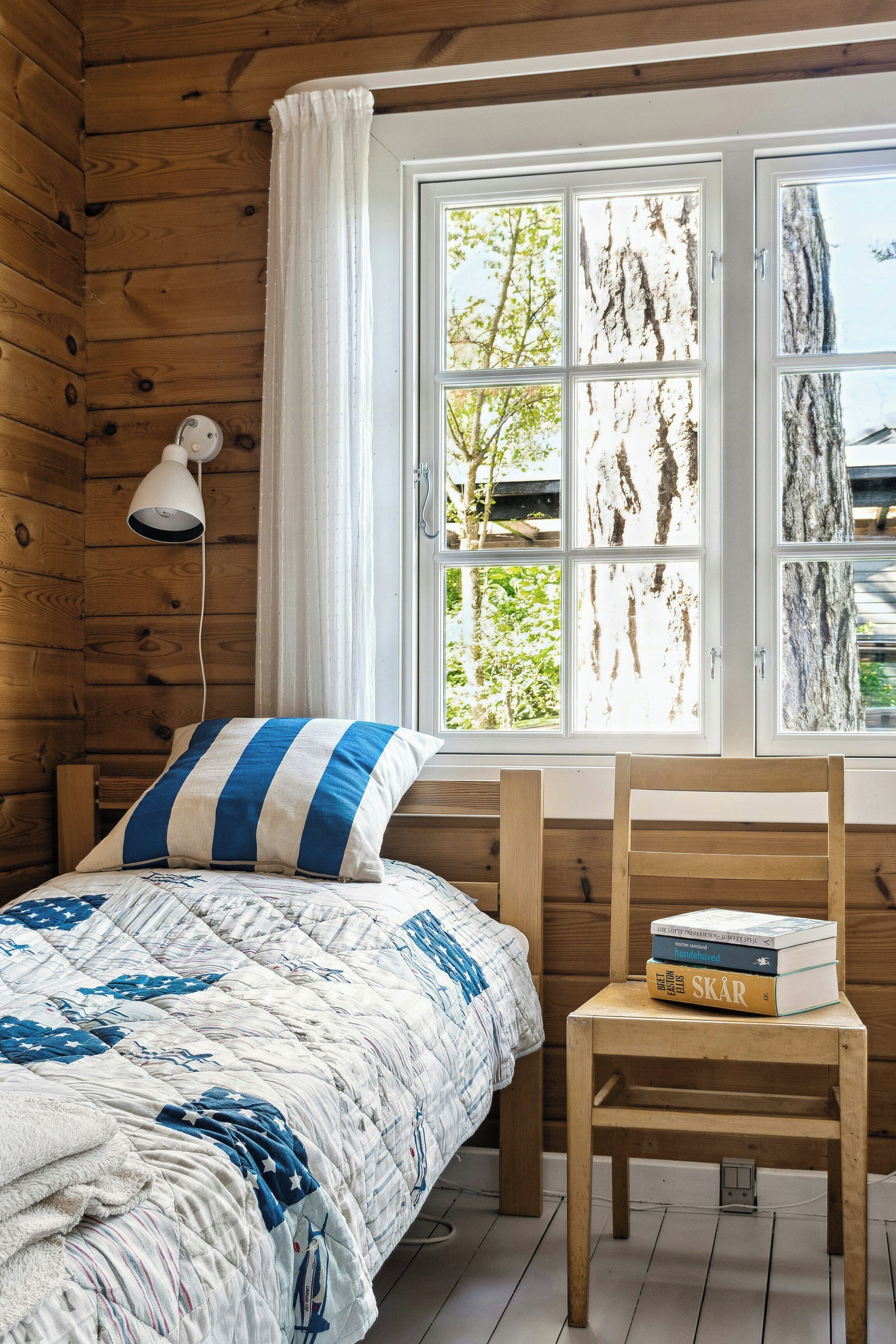 A wooden cabin room with a bed, a bedside lamp, a window with a view of trees, and a chair with books.