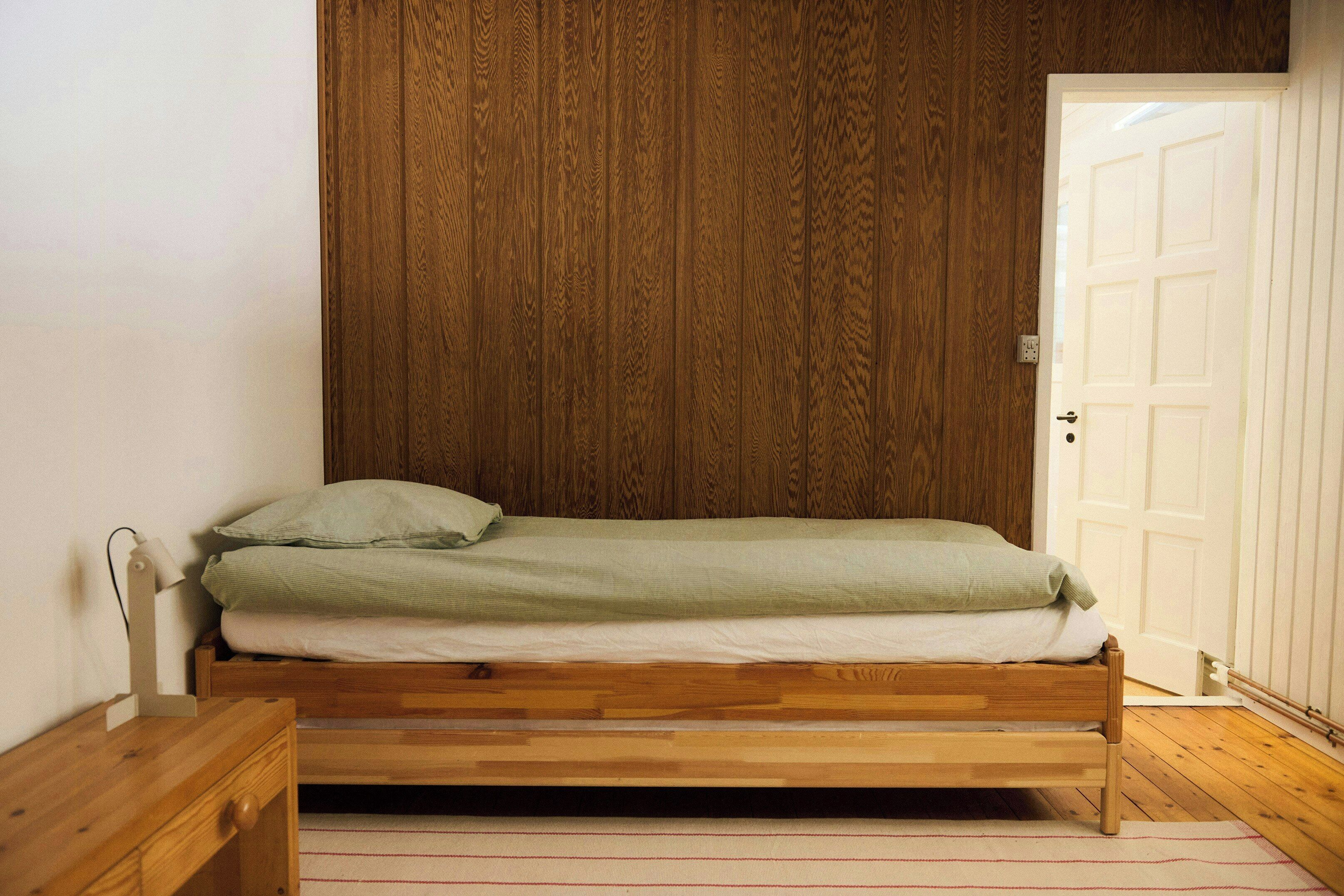 A wooden bed with a green mattress and pillows, a wooden nightstand with a lamp, and a white door in a room with wooden paneling on the wall.