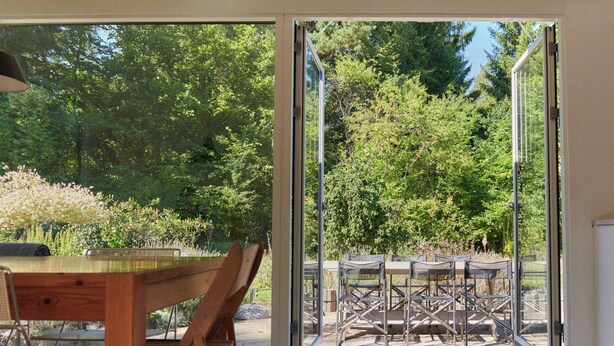 Une table et des chaises en bois se trouvent à l’intérieur d’une pièce. Une porte vitrée donne accès à un espace extérieur aménagé avec des tables et des chaises.
