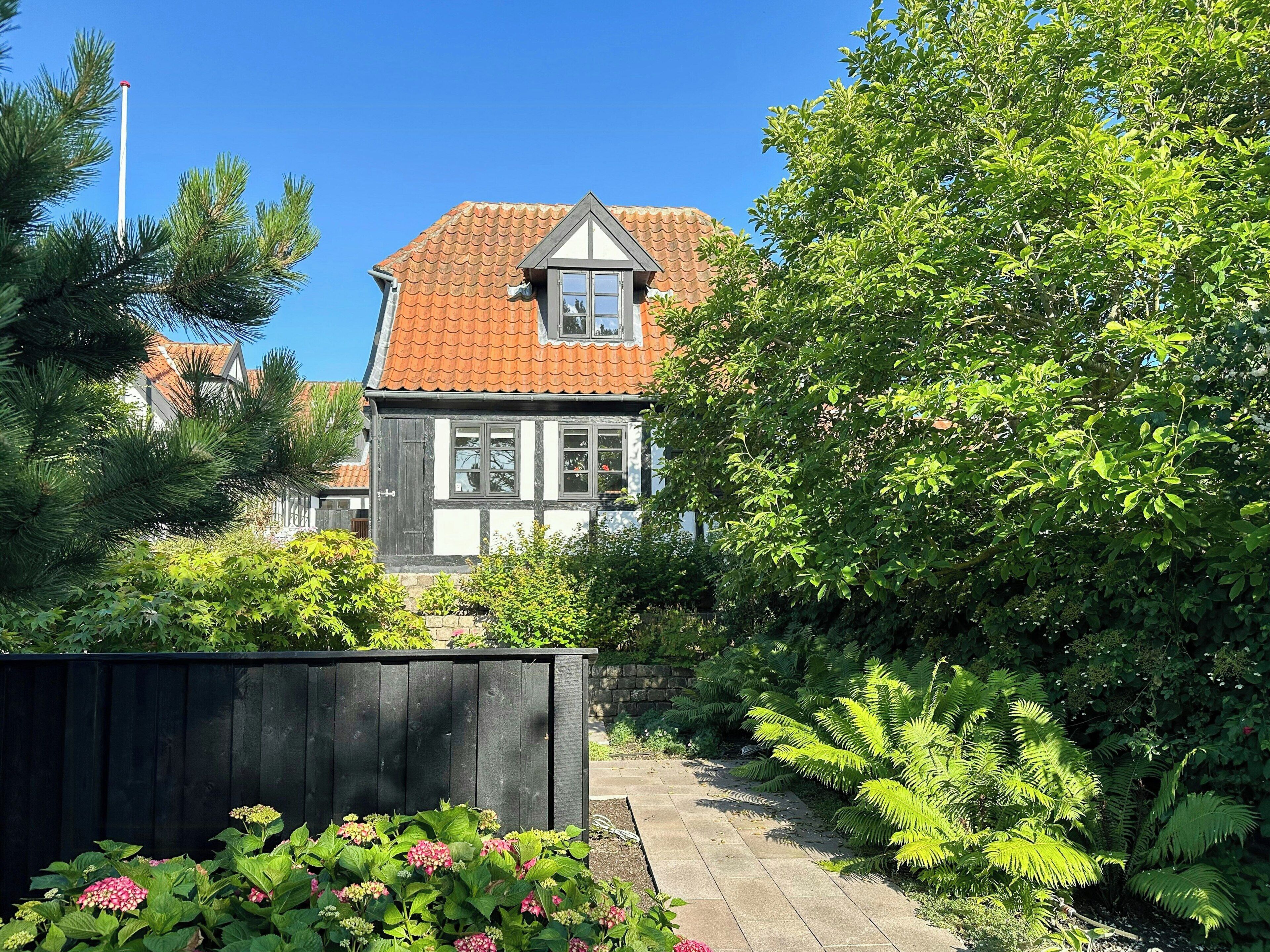 Une maison traditionnelle avec un toit en tuiles, entourée d’une végétation luxuriante et d’une clôture en bois.