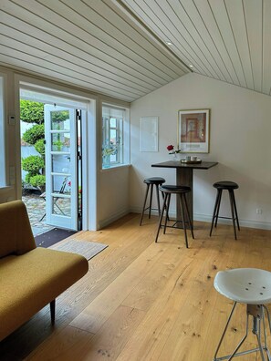 A room with a wooden floor, a sofa, a dining table with stools, a door leading outside, and a framed picture on the wall. - Half-timbered house with harbor view in Ebeltoft (Ebeltoft)