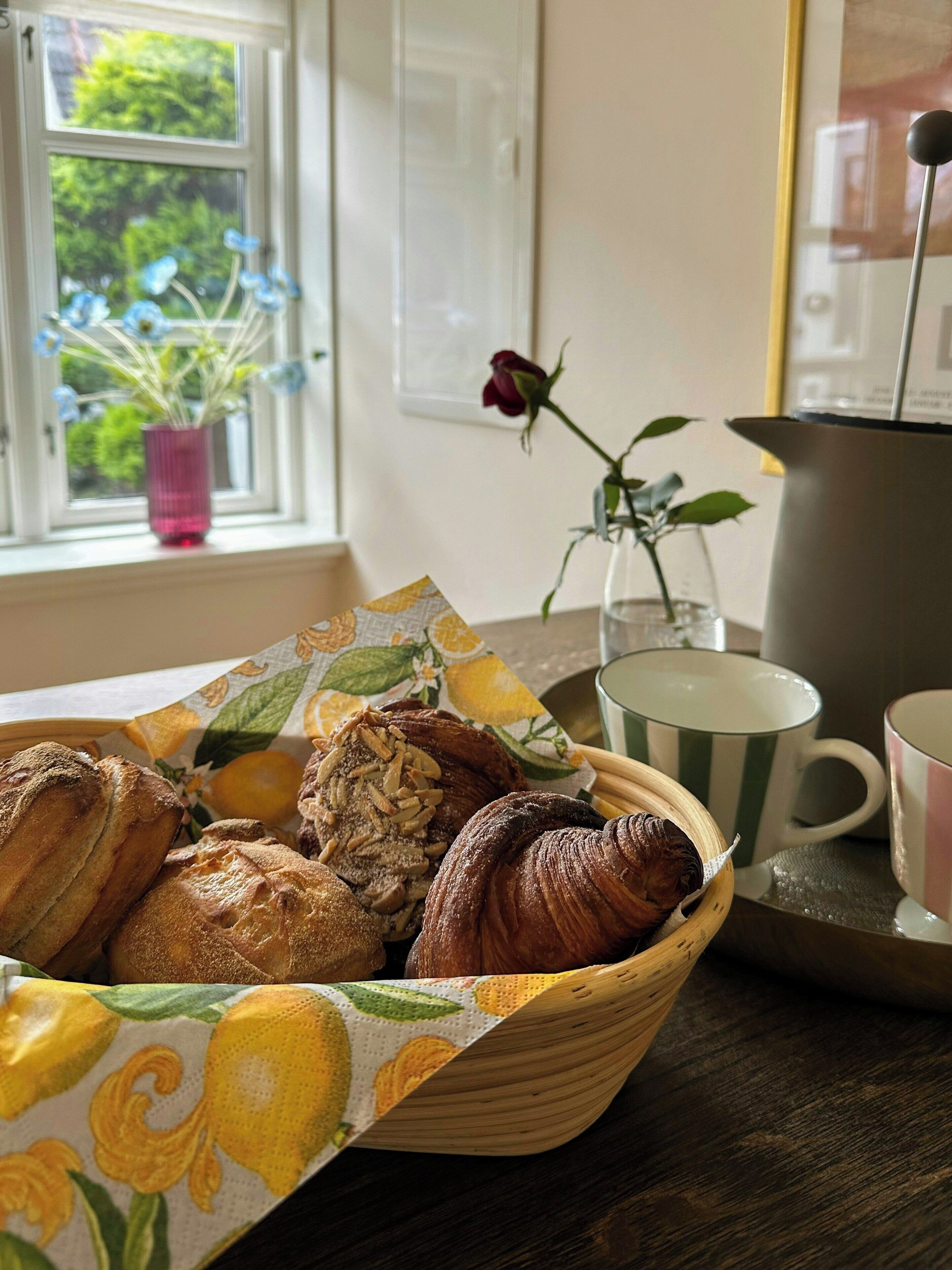 Un panier rempli de viennoiseries est posé sur une table près d’une fenêtre, à côté d’une plante en pot et d’un vase contenant une seule rose.
