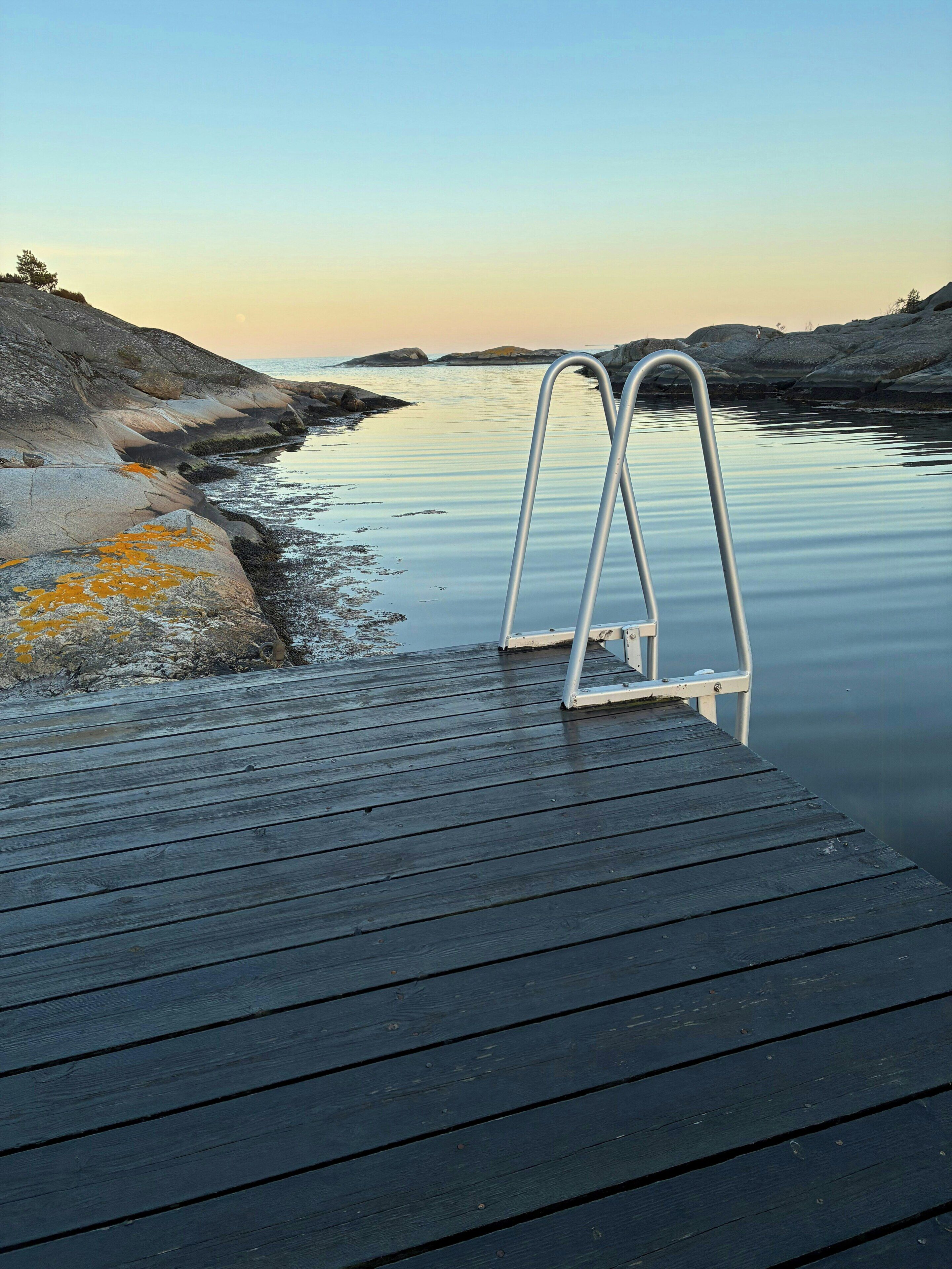 Un muelle de madera con una escalera metálica que desciende a aguas tranquilas, rodeado de terreno rocoso.