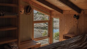 A wooden cabin interior with a bed, shelves, and a window overlooking rocky terrain.