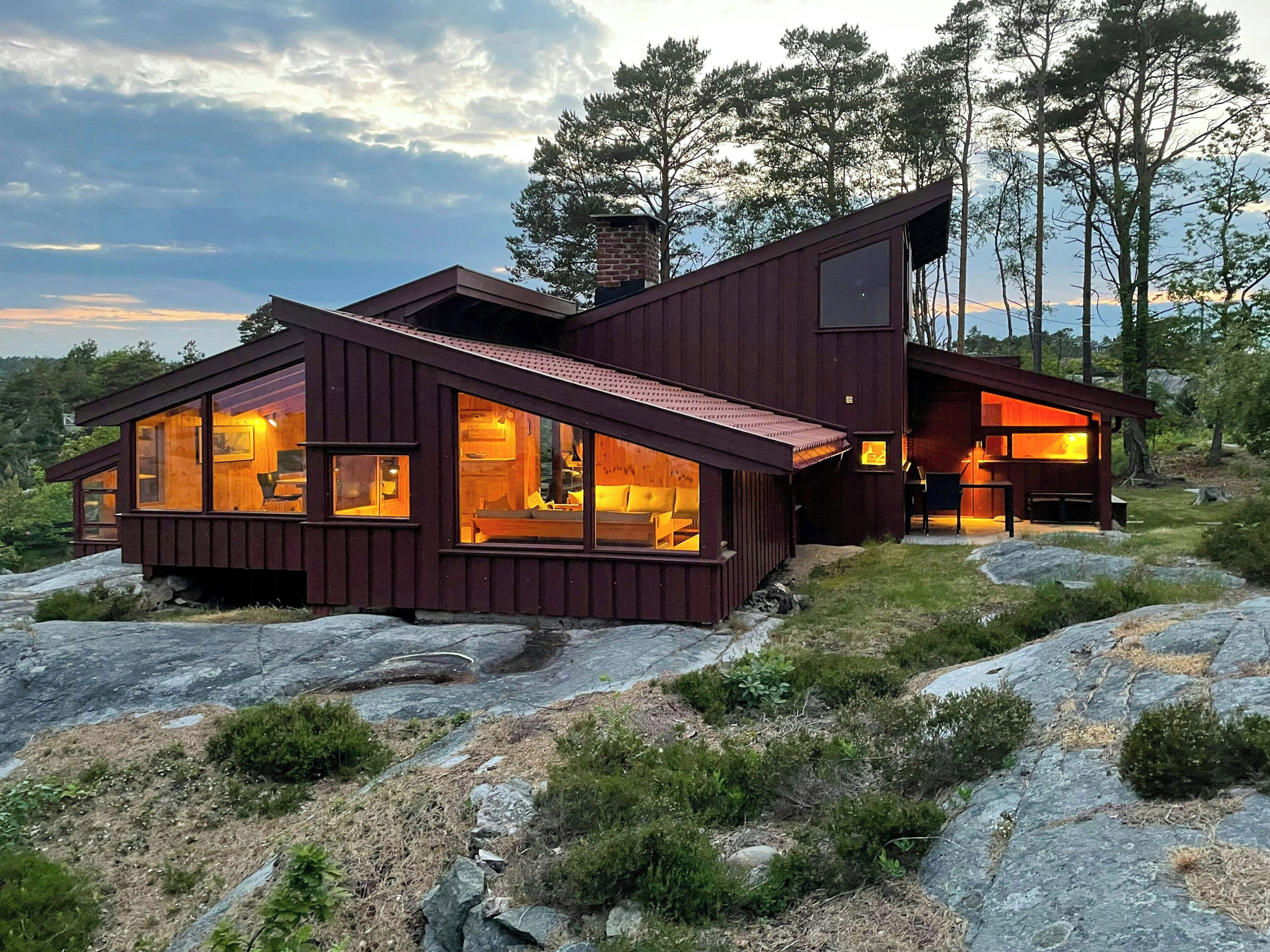A modern wooden cabin with large windows, situated on rocky terrain with trees in the background.