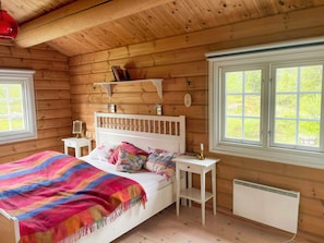 A wooden cabin bedroom with a bed, bedside tables, a heater, and a window.
