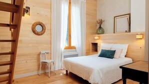 A bedroom with a wooden floor, a bed with white bedding, a chair, a window with white curtains, and a wall-mounted light fixture. - Eco-friendly gite in a wine estate in Frontignan (Frontignan)